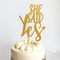 Gold 'She Said Yes' cake topper on a white cake with a white background