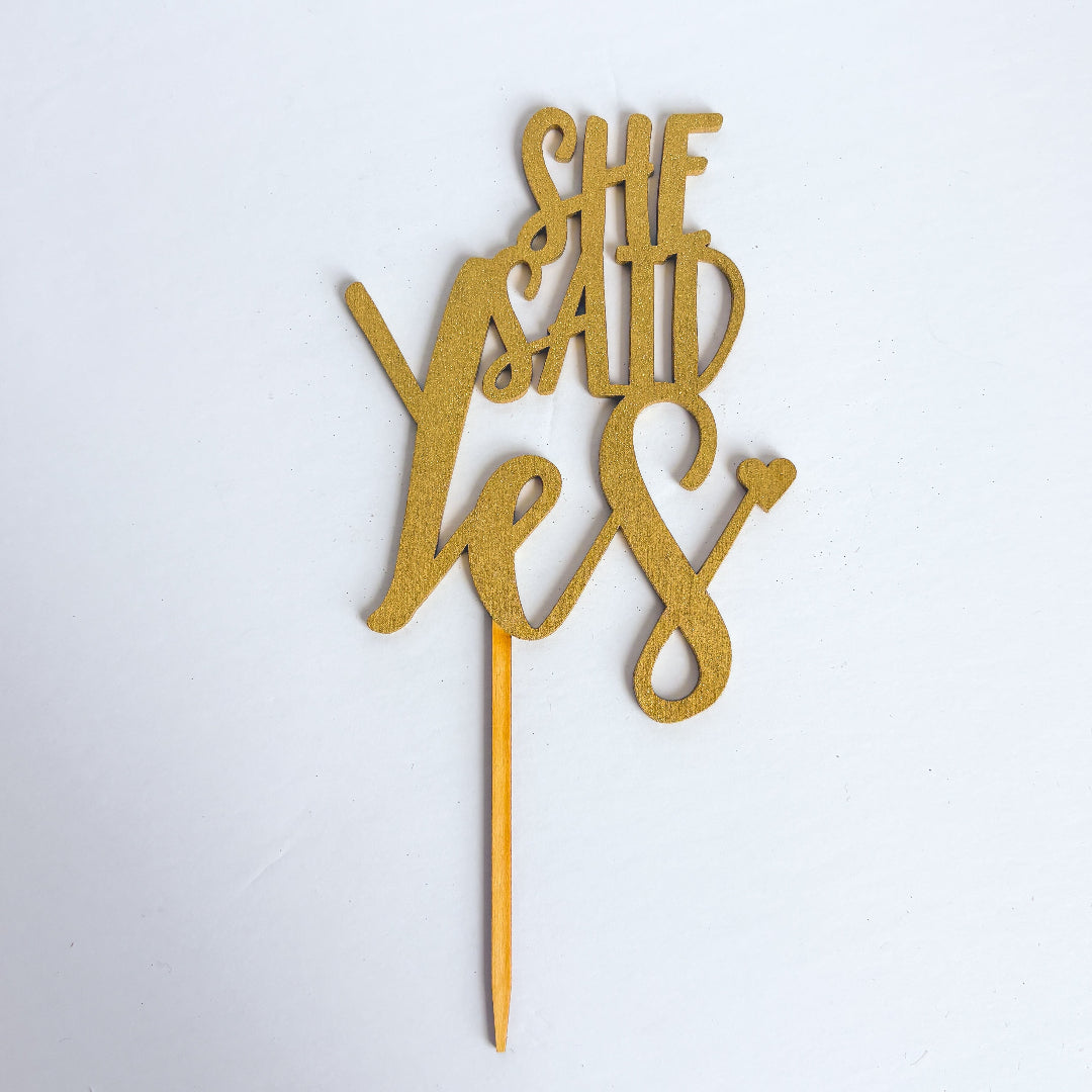 Gold 'She Said Yes' cake topper on a white background