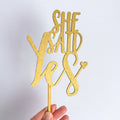 Gold glitter 'She Said Yes' cake topper held by a hand against a white background