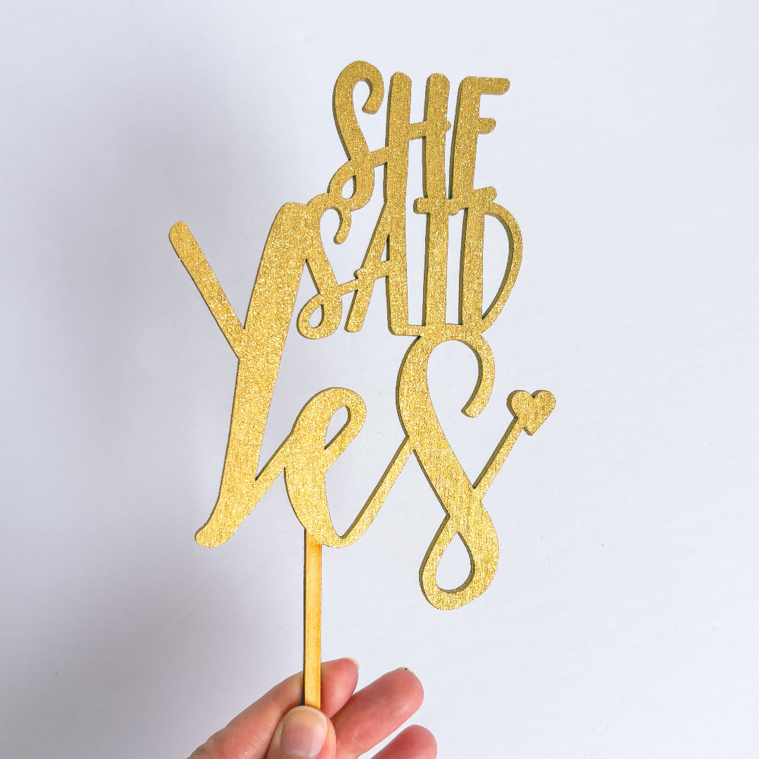 Gold glitter 'She Said Yes' cake topper held by a hand against a white background