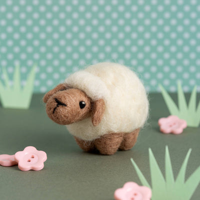 Small felt sheep toy on a green surface with pink flowers and green grass against a polka dot background