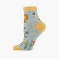 A pair of light blue ankle socks with yellow cuffs and toes. The socks feature an orange butterfly at the top and various small flowers and butterflies scattered across. Text on the socks reads "PLEASE, ONE SHIT STORM AT A TIME".