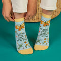 A pair of light blue ankle socks with yellow cuffs and toes. The socks feature an orange butterfly at the top and various small flowers and butterflies scattered across. Text on the socks reads "PLEASE, ONE SHIT STORM AT A TIME".