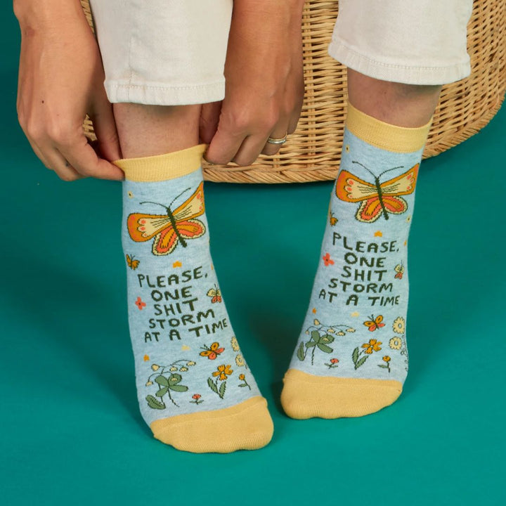 A pair of light blue ankle socks with yellow cuffs and toes. The socks feature an orange butterfly at the top and various small flowers and butterflies scattered across. Text on the socks reads 