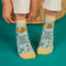 A pair of light blue ankle socks with yellow cuffs and toes. The socks feature an orange butterfly at the top and various small flowers and butterflies scattered across. Text on the socks reads 