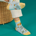 A pair of light blue ankle socks with yellow cuffs and toes. The socks feature an orange butterfly at the top and various small flowers and butterflies scattered across. Text on the socks reads "PLEASE, ONE SHIT STORM AT A TIME".