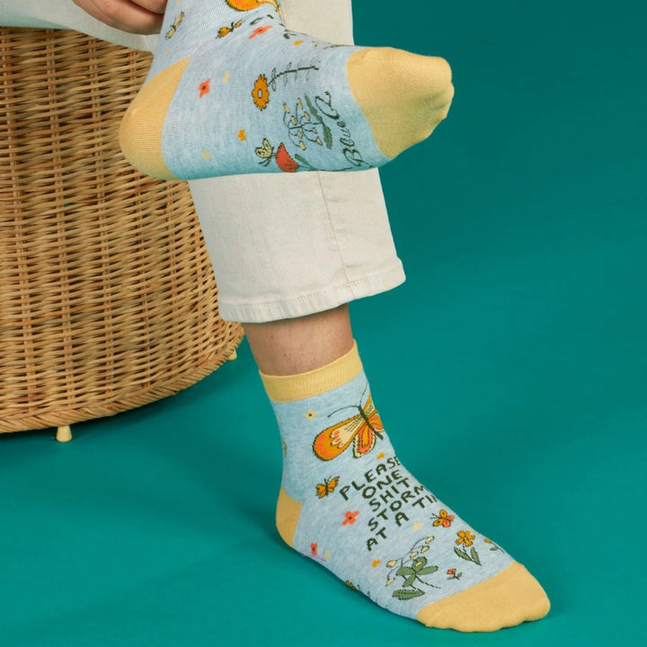 A pair of light blue ankle socks with yellow cuffs and toes. The socks feature an orange butterfly at the top and various small flowers and butterflies scattered across. Text on the socks reads 