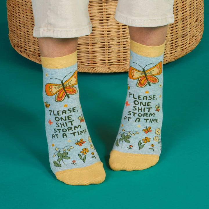 A pair of light blue ankle socks with yellow cuffs and toes. The socks feature an orange butterfly at the top and various small flowers and butterflies scattered across. Text on the socks reads 
