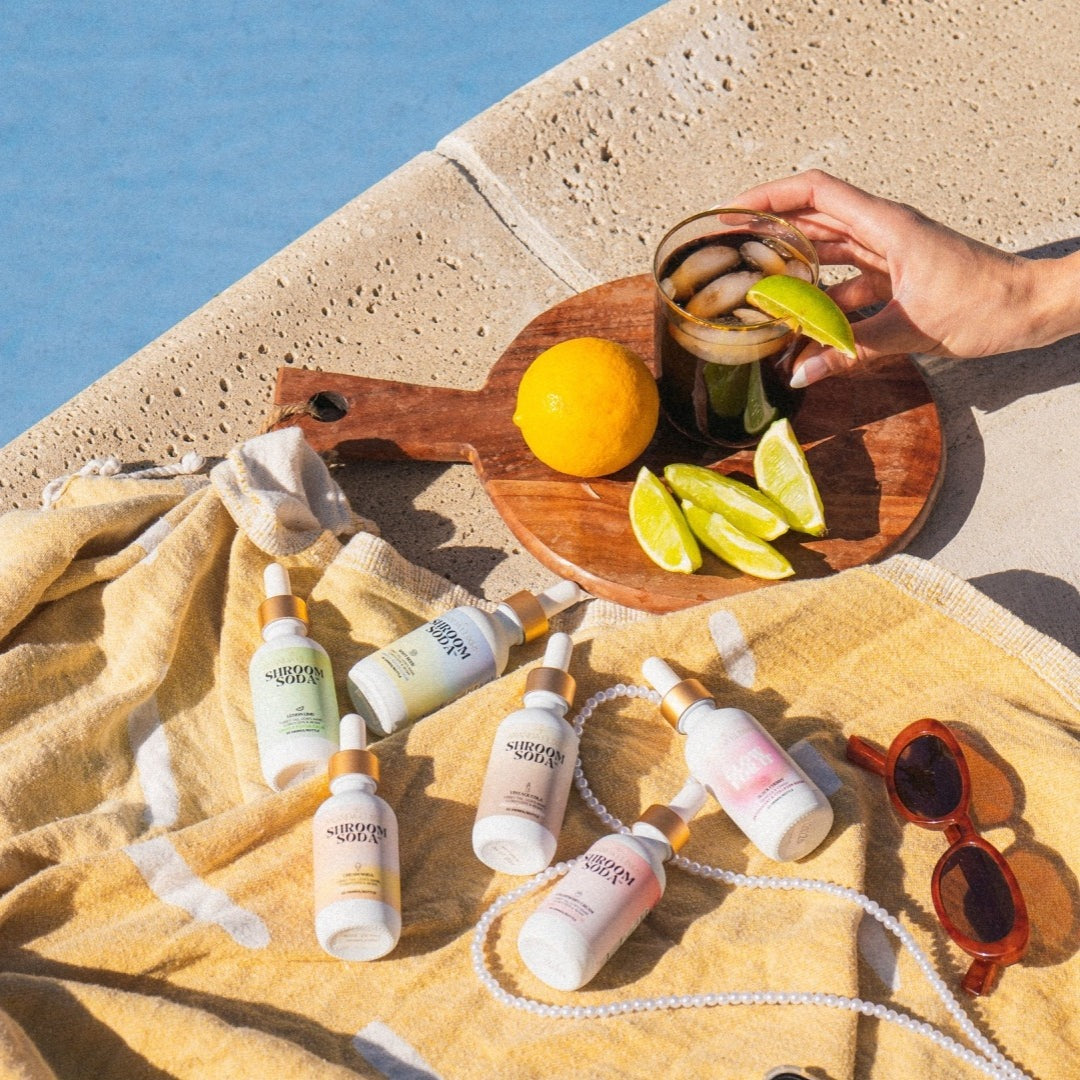 Sunscreen bottles on a towel with a wooden cutting board, lemon, lime, and sunglasses by the sea.