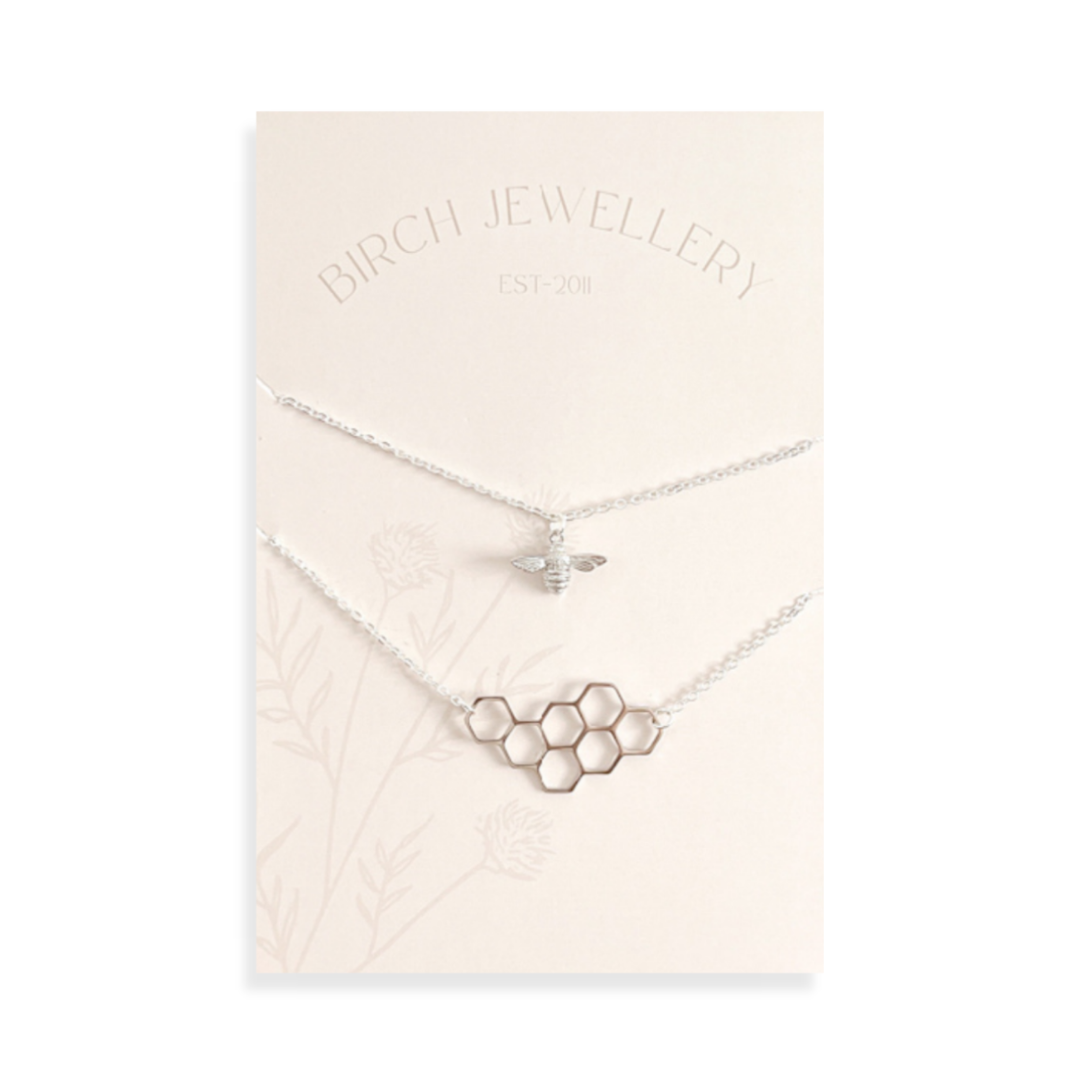 A pair of silver necklaces on a white card. The longer necklace features a honeycomb pendant, and the shorter necklace features a bee pendant. The card has the text "BIRCH JEWELLERY" and "EST-2011" printed on it.