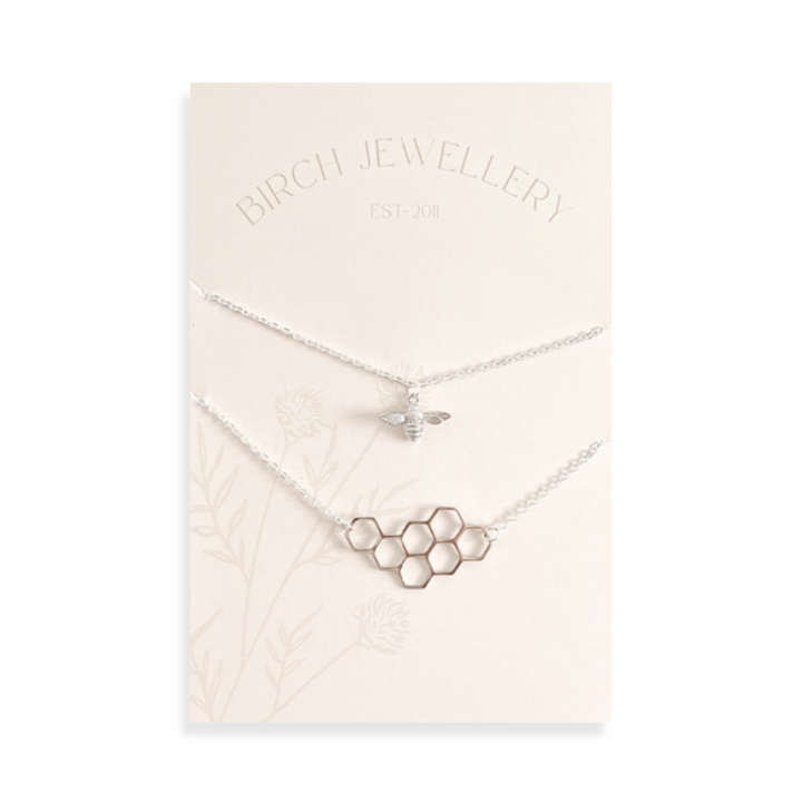 A pair of silver necklaces on a white card. The longer necklace features a honeycomb pendant, and the shorter necklace features a bee pendant. The card has the text 