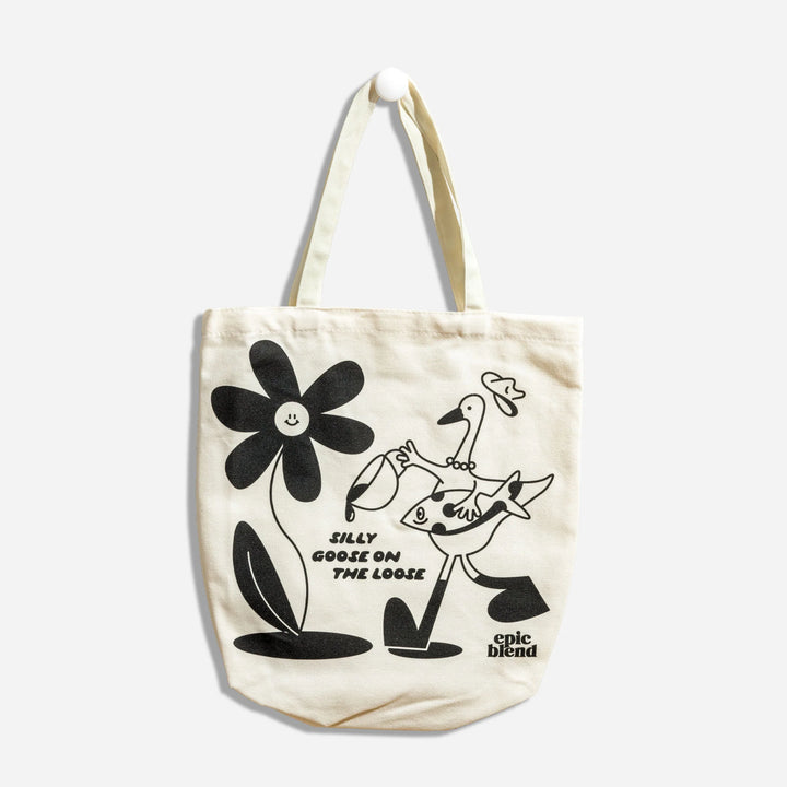 A white tote bag with a black and white illustration. The illustration shows a goose pouring coffee on a flower with a smiley face. The text 