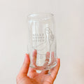 A hand holding a clear glass can-shaped tumbler with a frosted etched design of a goose holding a beer glass and the text 'SILLY GOOSE JUICE' against a plain light background.