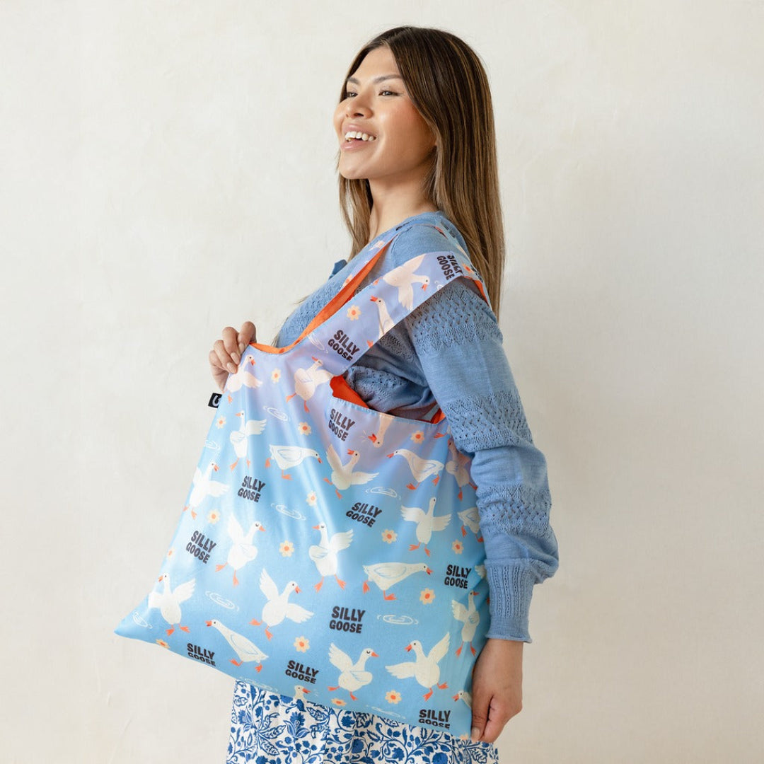 A woman wearing a blue knit sweater and patterned skirt holds a light blue reusable tote bag printed with white silky geese and orange handles against a plain off-white background.