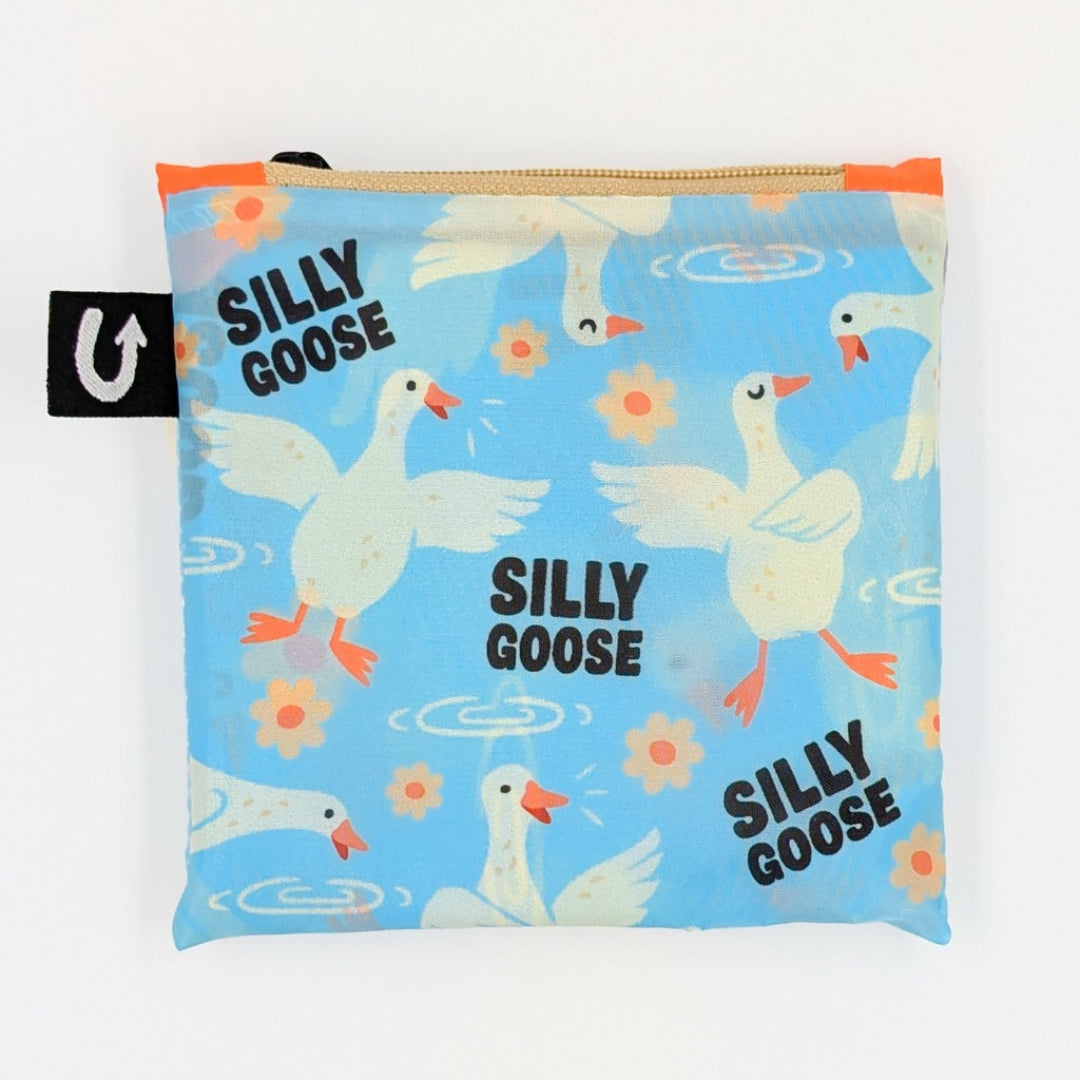A square zippered pouch with a light blue background printed with white geese, orange beaks and feet, and the words 'SILLY GOOSE' repeated. The pouch has a black fabric tag with a white U logo on the left side and a gold zipper at the top.
