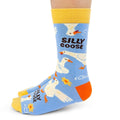 A pair of crew socks worn on a lower leg, featuring a light blue background with cartoon white geese, orange beaks and feet, yellow cuffs and toes, and the words 'SILLY GOOSE' printed on the front.