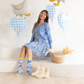 A woman sits on a white platform in a bright studio setting, wearing a blue patterned dress, blue cardigan, and colorful socks. Two white goose-shaped balloons and blue-and-white gingham heart balloons float behind her, with a wicker basket and cream-colored boots on the floor nearby.