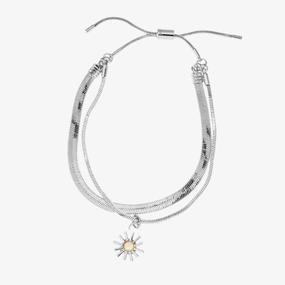 A set of two silver bracelets, one layered over the other, with a sun charm hanging from the bottom. The bracelets are displayed on a pink card with the brand name 