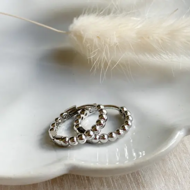 A pair of 925 sterling silver ball hoop earrings, featuring a beaded design, displayed on a white textured surface with white floral accents. The earrings are dainty and high-quality, available in Ottawa, Ontario, Canada.