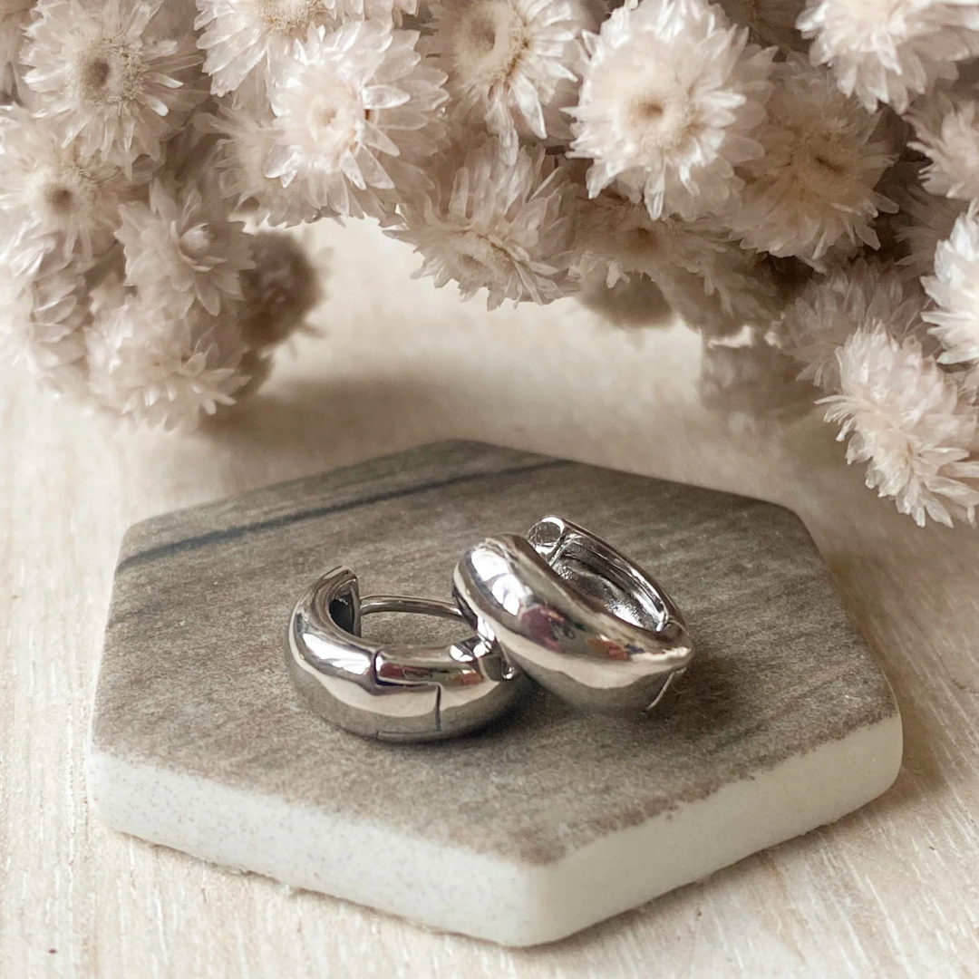 A pair of silver hoop earrings displayed on a marble-like surface with a bouquet of dried white flowers in the background.