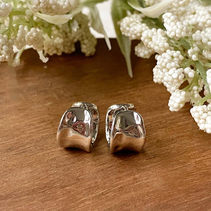 A pair of gold Grace hoops, featuring a chunky, domed design, displayed on a textured wooden surface with white floral accents. The earrings are statement huggie hoops, available in Ottawa, Ontario, Canada.