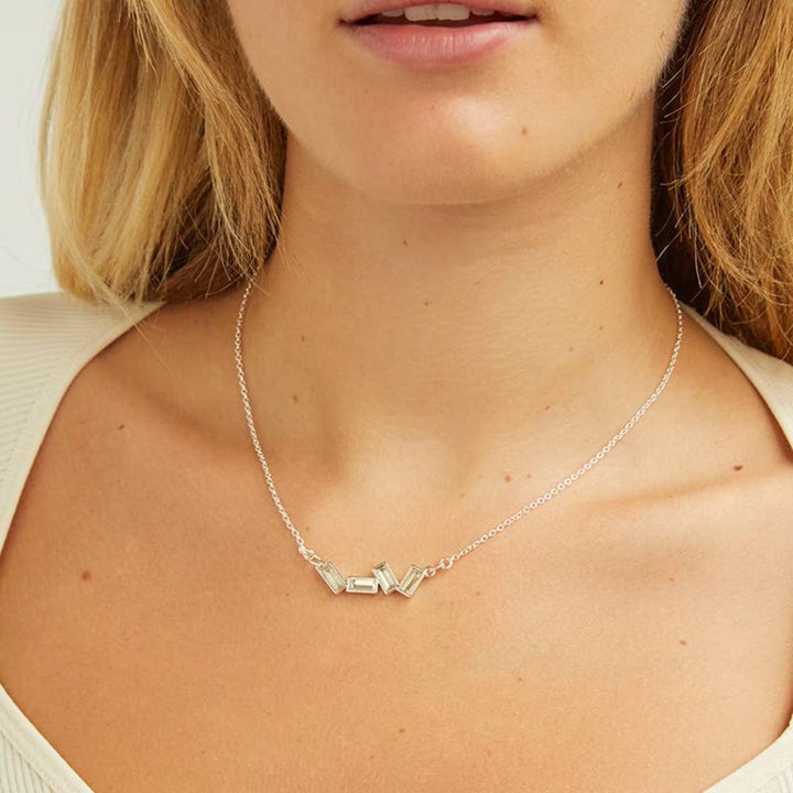 A silver necklace with several rectangular-shaped crystals on a pink card with the brand name 