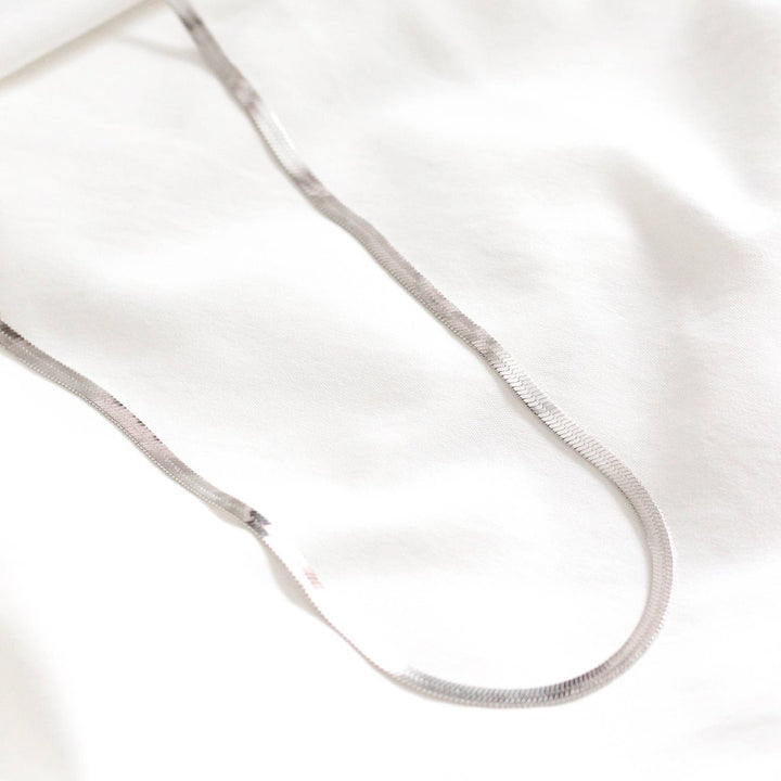 A silver herringbone chain necklace resting on a white surface. The chain is flat and has a wavy, snake-like pattern.