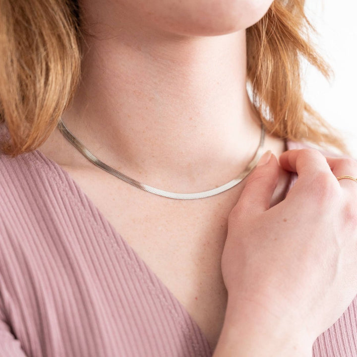 A silver herringbone chain necklace resting on a white surface. The chain is flat and has a wavy, snake-like pattern.