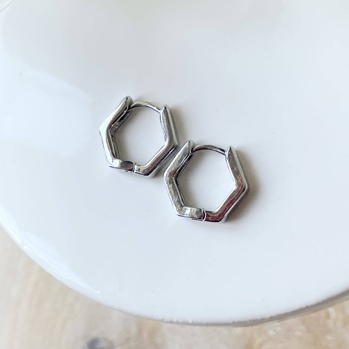 A pair of silver Hexi huggies, featuring a minimalist, hexagon hoop design, displayed on a white ceramic dish with white floral accents. The earrings are geometric and modern, available in Ottawa, Ontario, Canada.