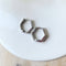 A pair of silver Hexi huggies, featuring a minimalist, hexagon hoop design, displayed on a white ceramic dish with white floral accents. The earrings are geometric and modern, available in Ottawa, Ontario, Canada.