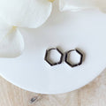 A pair of silver Hexi huggies, featuring a minimalist, hexagon hoop design, displayed on a white ceramic dish with white floral accents. The earrings are geometric and modern, available in Ottawa, Ontario, Canada.