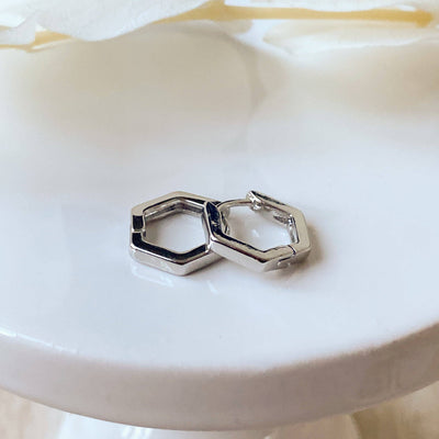 A pair of silver Hexi huggies, featuring a minimalist, hexagon hoop design, displayed on a white ceramic dish with white floral accents. The earrings are geometric and modern, available in Ottawa, Ontario, Canada.