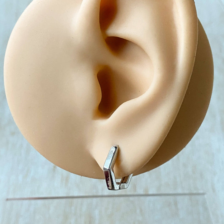 A pair of silver Hexi huggies, featuring a minimalist, hexagon hoop design, displayed on a white ceramic dish with white floral accents. The earrings are geometric and modern, available in Ottawa, Ontario, Canada.