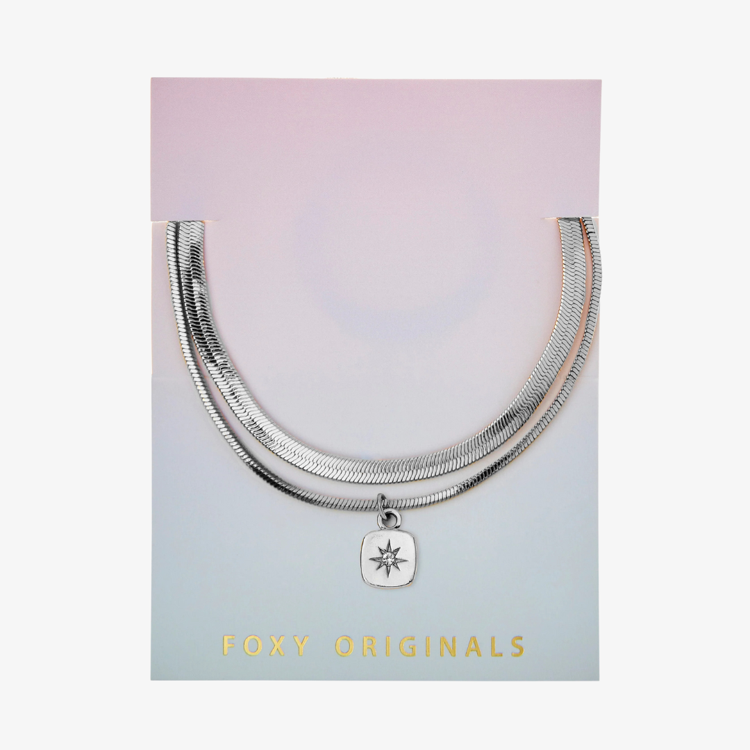 A set of two silver bracelets, one layered over the other, with a star charm hanging from the bottom. The bracelets are displayed on a pink card with the brand name "Foxy Originals."