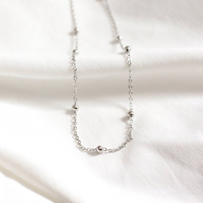A silver chain necklace with multiple small, round beads evenly spaced along the chain. The necklace is resting on a white surface.