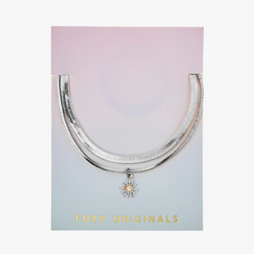 A set of two silver bracelets, one layered over the other, with a sun charm hanging from the bottom. The bracelets are displayed on a pink card with the brand name "Foxy Originals."