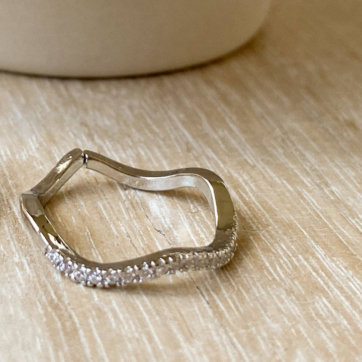 A silver Tidal Wave ring, featuring a wavy, curved design with pave diamonds, displayed on a light wooden surface with a blurred white ceramic element in the background. The ring is a curved wedding band or stacking ring, available in Ottawa, Ontario, Canada.
