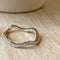 A silver Tidal Wave ring, featuring a wavy, curved design with pave diamonds, displayed on a light wooden surface with a blurred white ceramic element in the background. The ring is a curved wedding band or stacking ring, available in Ottawa, Ontario, Canada.