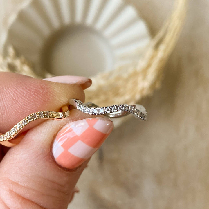A silver Tidal Wave ring, featuring a wavy, curved design with pave diamonds, displayed on a light wooden surface with a blurred white ceramic element in the background. The ring is a curved wedding band or stacking ring, available in Ottawa, Ontario, Canada.