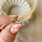 A silver Tidal Wave ring, featuring a wavy, curved design with pave diamonds, displayed on a light wooden surface with a blurred white ceramic element in the background. The ring is a curved wedding band or stacking ring, available in Ottawa, Ontario, Canada.