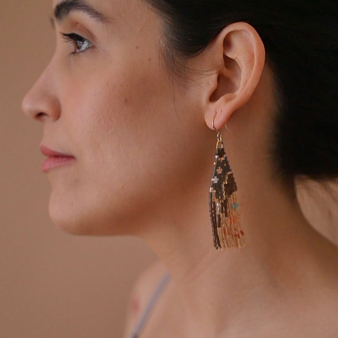 A pair of long, triangular tapestry-style earrings hand-stitched with small Japanese glass beads to depict a stylized face with light peach skin, blue eyes, coral cheeks, and a red mouth, framed by dark brown and grey hair with gold bead accents. They hang from delicate 14 KT gold filled fishhook ear wires.