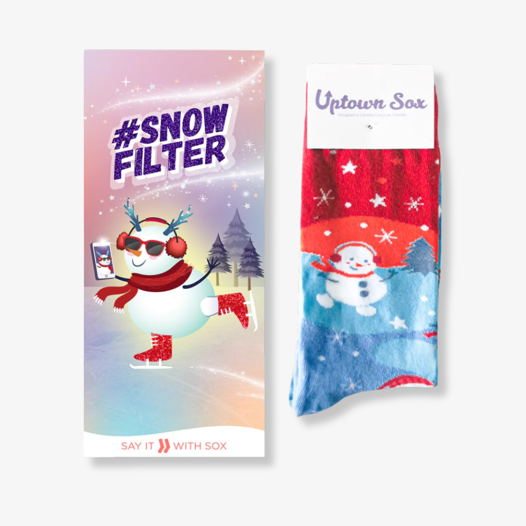A pair of colorful socks with a winter theme, packaged with a card. The socks feature a snowman ice skating and taking a selfie, with the hashtag "#SNOW FILTER" printed on the card.