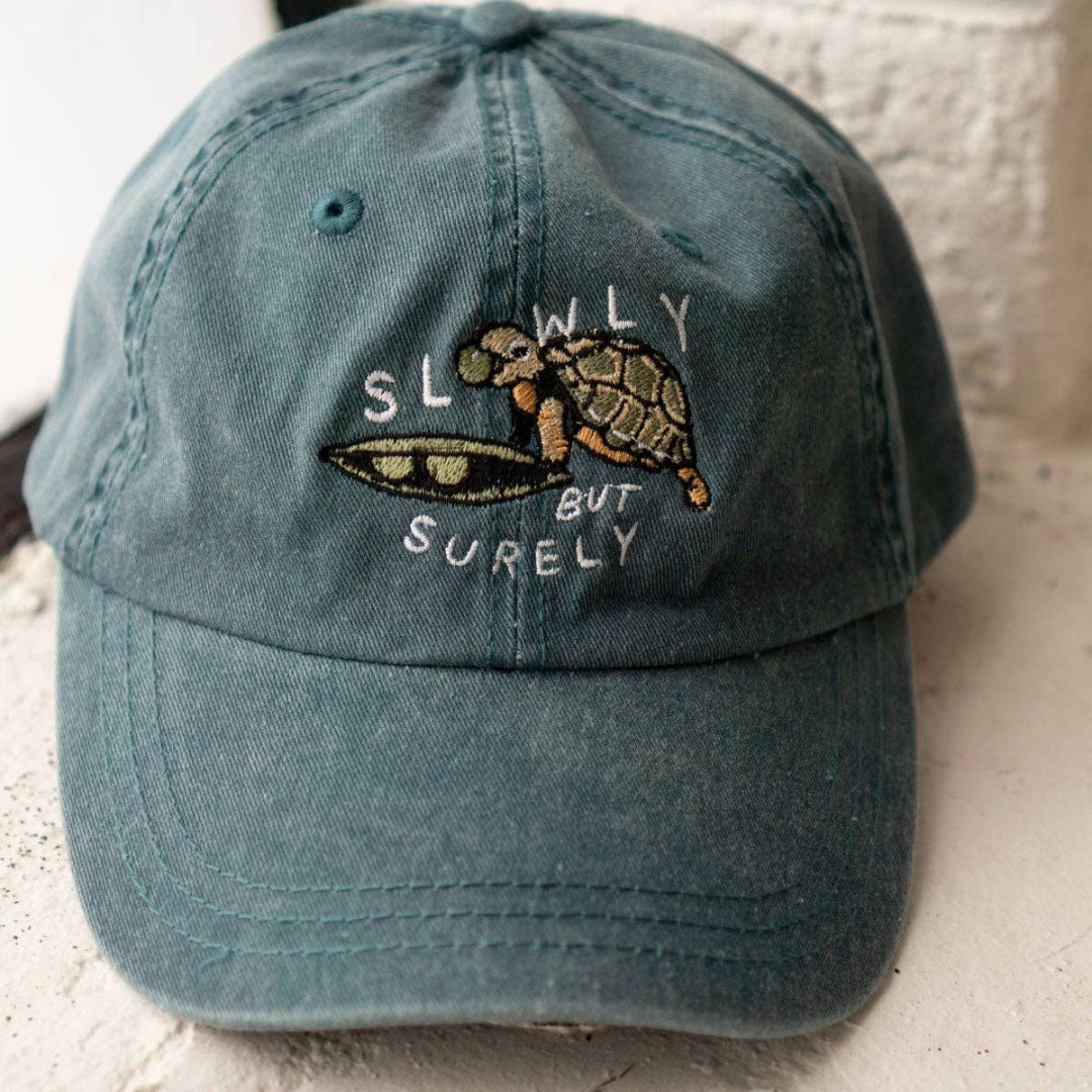 Blue cap with turtle design and 'Slowly but Surely' text on a white surface.