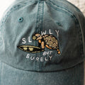 Blue cap with turtle design and text 'SLOWLY BUT Surely' on a blurred background