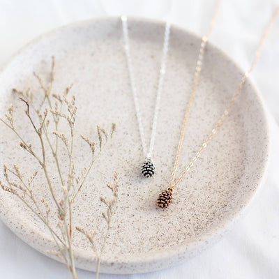 Two pinecone necklaces on a textured stone surface with dried plants.