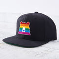 A black snapback cap with a flat green brim. The front features a unique bear face patch in the colours of the rainbow pride flag. Shop this at Cheerfully Made, Canadian-made gift shop.