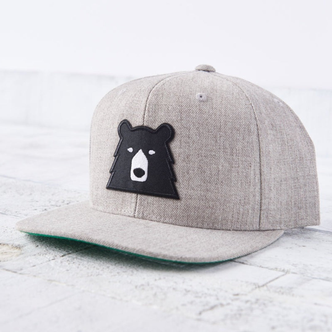 A heather grey snapback cap with a flat green brim. The front features a black bear face patch with white eyes and snout. Shop this at Cheerfully Made, Canadian-made gift shop.
