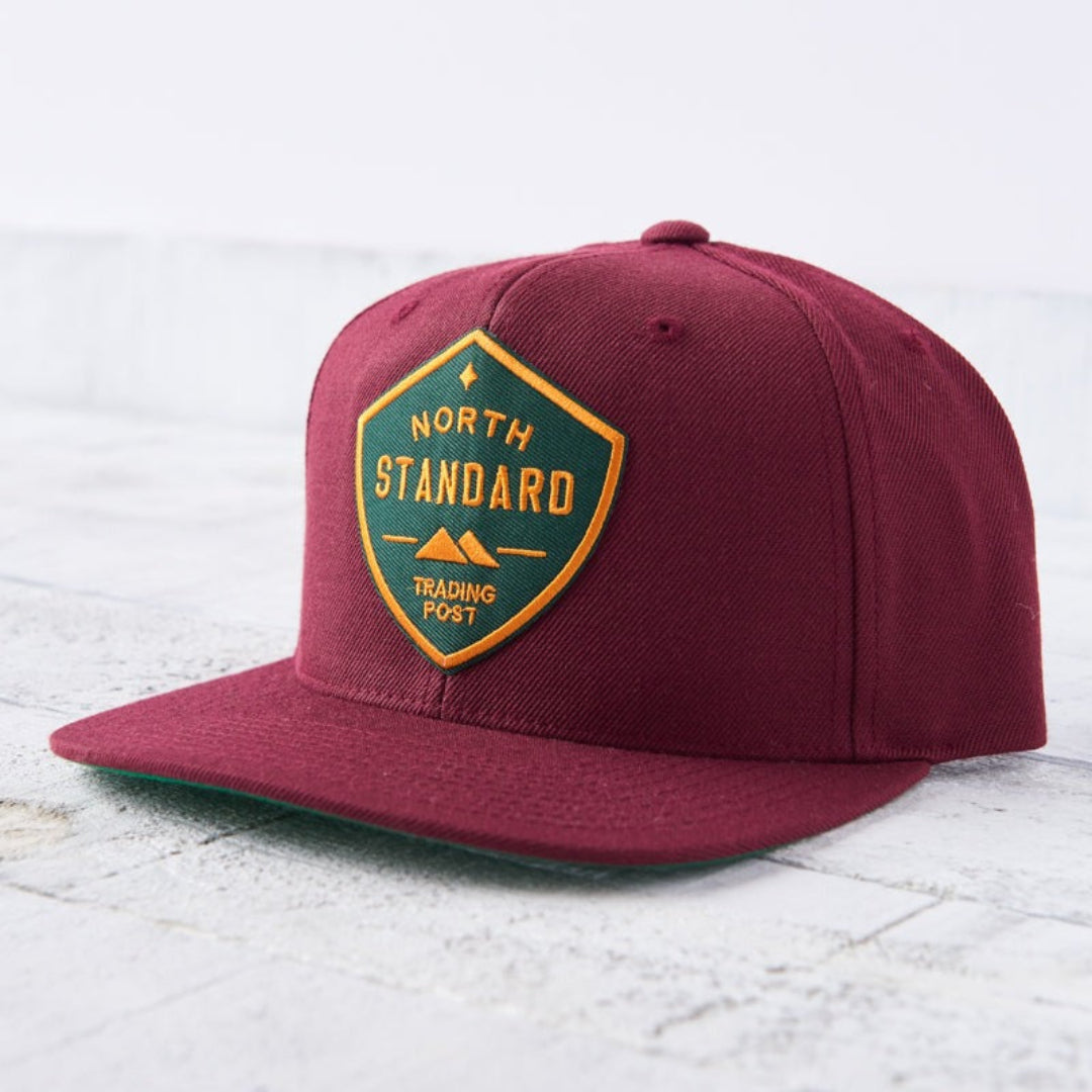 A maroon-coloured snapback cap with a flat green brim. The front features a green and gold shield-shaped patch with "NORTH STANDARD TRADING POST" text and a small mountain graphic. Shop this at Cheerfully Made, Canadian-made gift shop.
