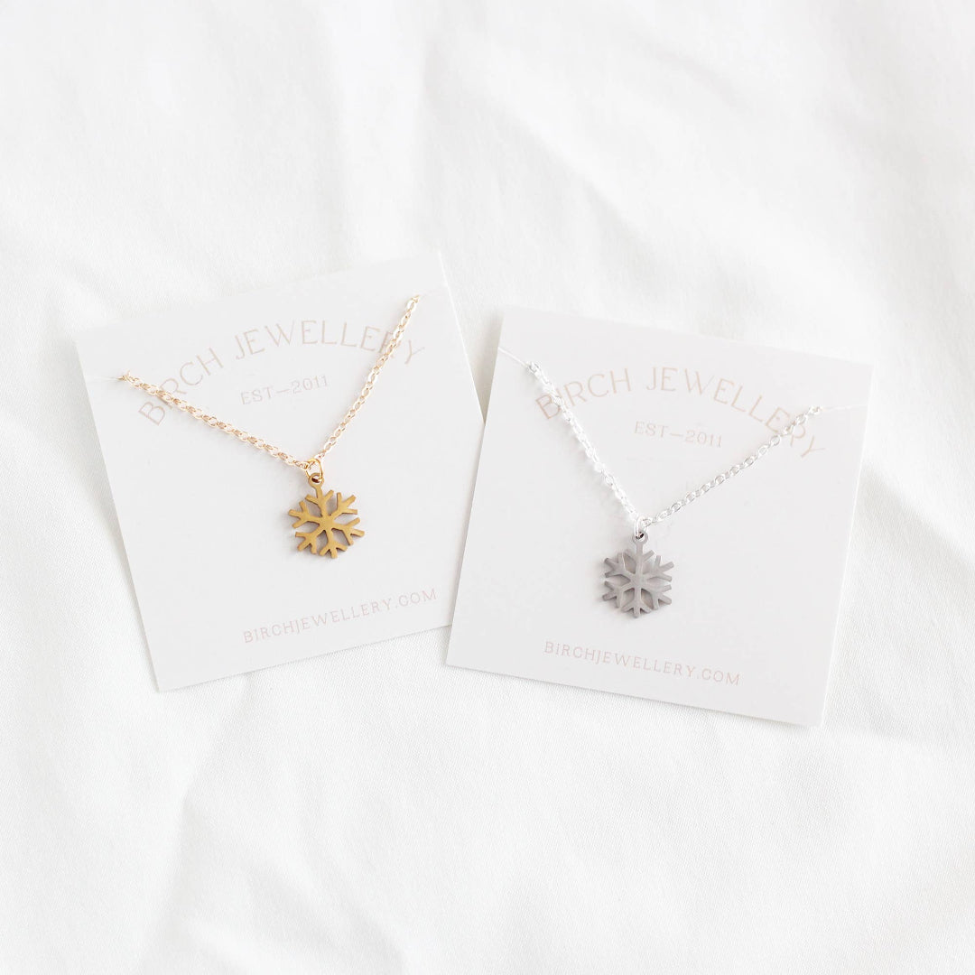 Two snowflake necklaces, one gold and one silver, on their respective cards with 'ARCH JEWELLERY' branding.