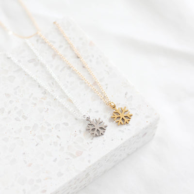 Two snowflake-shaped pendants on gold necklaces against a white textured background